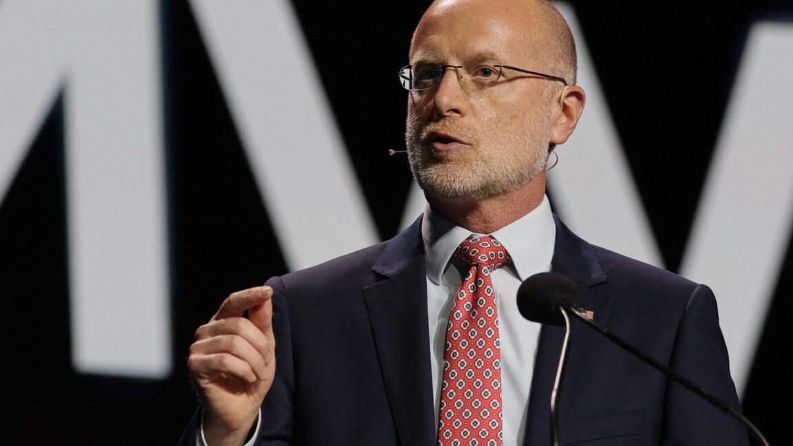FCC Chairman Brendan Carr delivers a speech on a stage while standing in front of a microphone and gesturing with his hand.