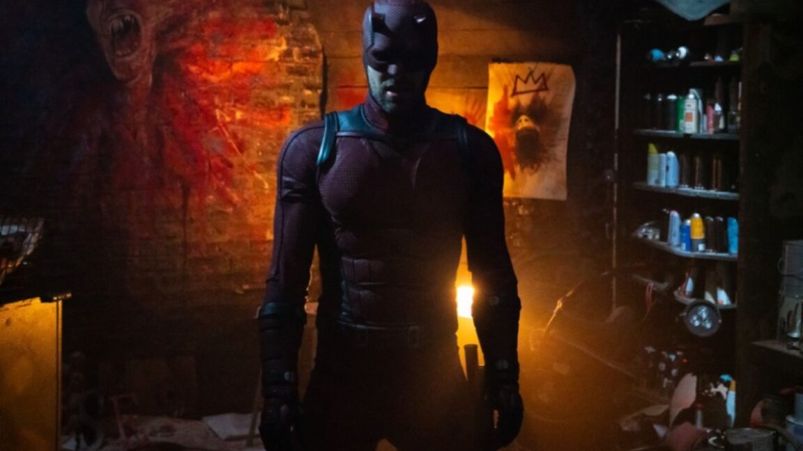 man in daredevil suit swathed in red light as he stands in a room with exposed brick walls