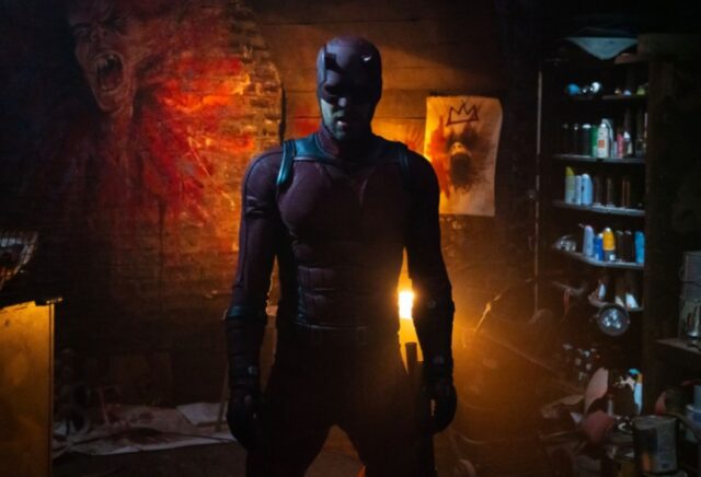 man in daredevil suit swathed in red light as he stands in a room with exposed brick walls