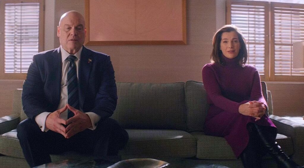 heavyset bald man in a dark suit and a pretty brunette woman in a red sweater seated on opposite ends of a couch.