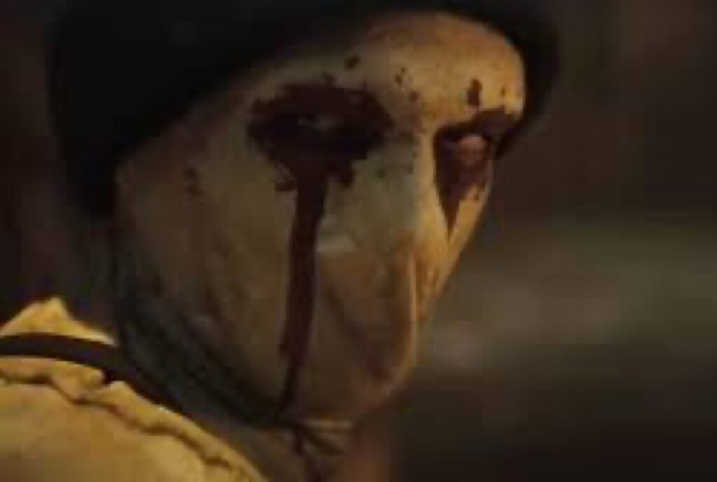 close-up of a man in a dirty white face mask with blood painting rivulets running down his face from his eyes.