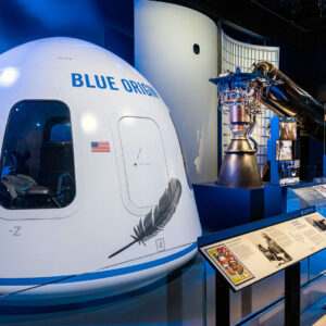 a gumdrop-shape white space capsule is seen on display with other rocket hardware in a museum gallery with blue walls and flooring