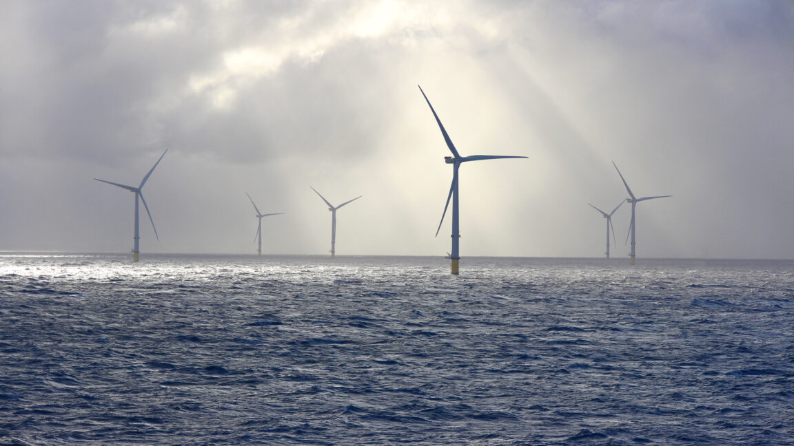 A series of tall wind turbines in the ocean, illuminated by beams of sunlight filtering through baps of heavy clouds.