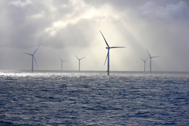 A series of tall wind turbines in the ocean, illuminated by beams of sunlight filtering through baps of heavy clouds.