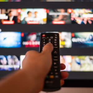 Hand Using A Television Remote Control in front of a TV with a streaming app on display and blurred in the background.