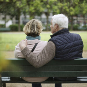 An elderly couple sitting on a public bench in a public garden.