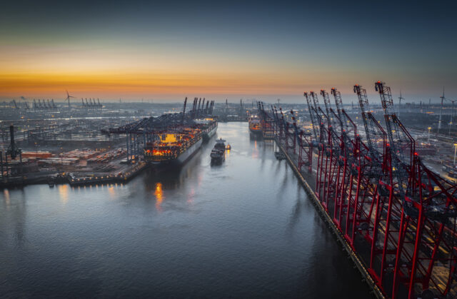 Aerial view of a container terminal with large container cargo ships and gantry cranes. A cargo ship is navigating through the central waterway. On both sides of the waterway are massive container terminals lined with red and black gantry cranes used for loading and unloading ships. on the left docked ships with brightly lit containers. Moody sky at dawn.