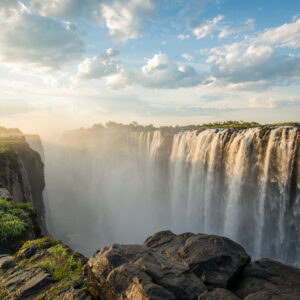 a view of a broad waterfall, with mist rising above rocks in the foreground.
