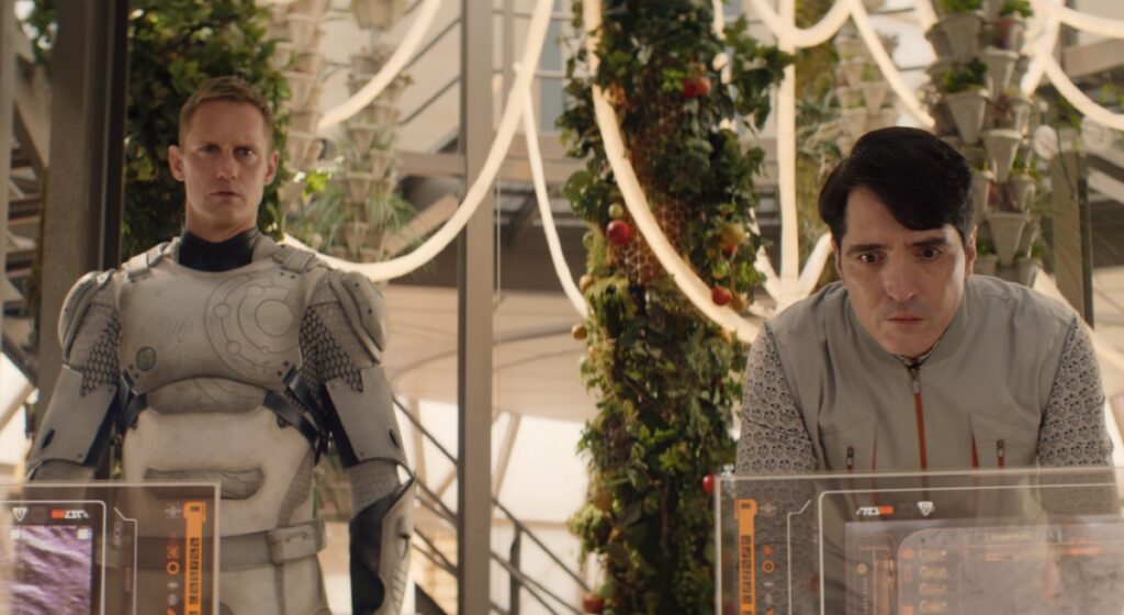 Murderbot with human face reveled standing at attention while a scientist leans over a computer monitor