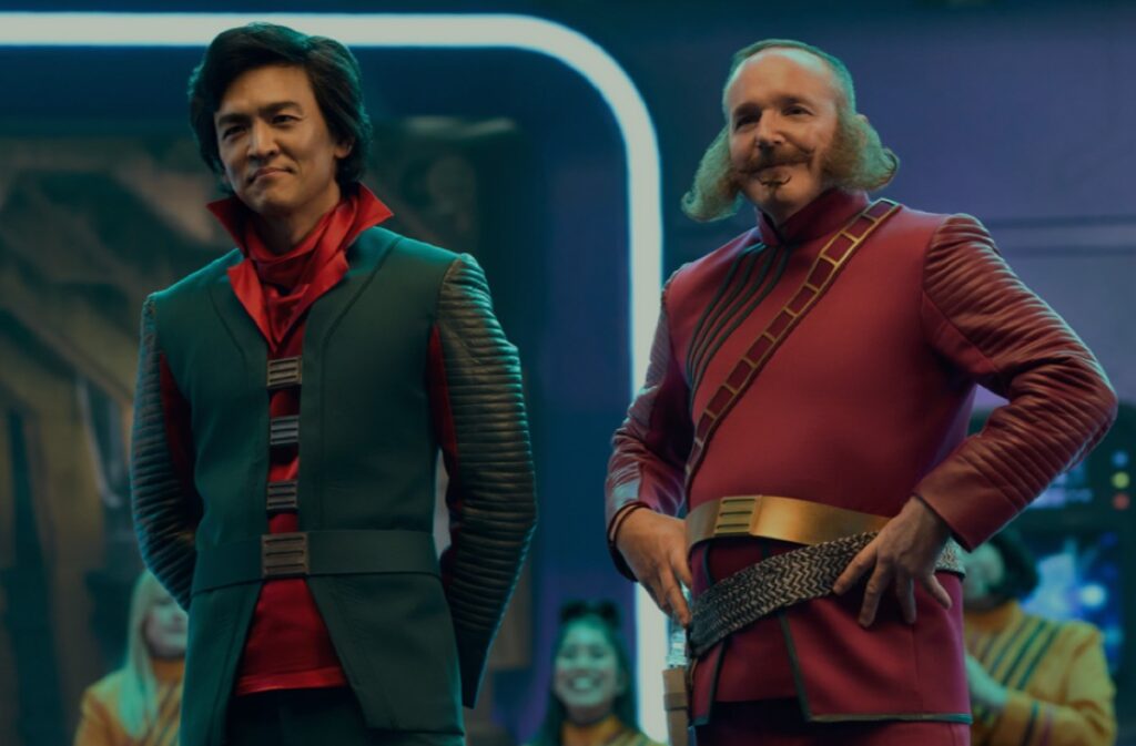 asian man in green jacket and red shirt, standing next to older man with sideburns in a red tunic with gold belt.