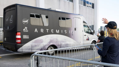 A motor coach decorated with an image of the moon is parked outside of a building.