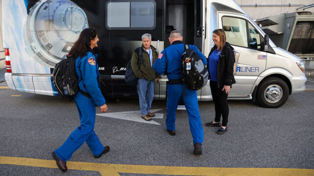 A woman and a man in blue flights join another woman and man as they board a motor coach with a spacecraft-themed graphic wrap.
