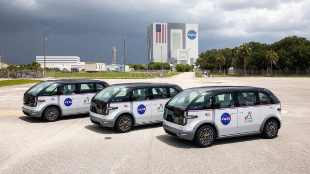 Three vans with rounded corners are parked side by side in front of a large building and an overcast sky.
