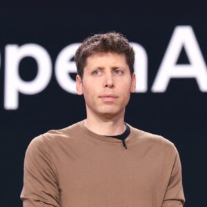 OpenAI CEO Sam Altman standing in front of a backup with a large OpenAI logo.