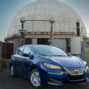 A blue Nissan Leaf in front of some electrical equipment and what looks like a concrete dome