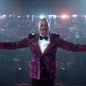 black man in colorful purple suit with arms outstretched in front of a camera with huge crowd behind him
