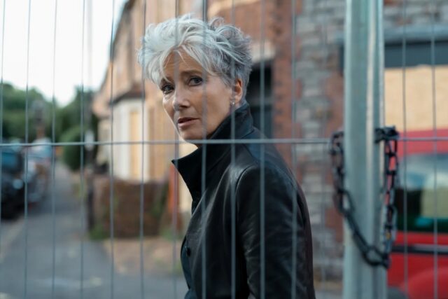 short gray-haired room in black coat staring through a mesh fence